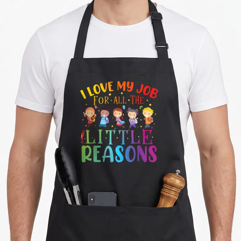 Reason teaching apron - Five cartoon children running under I LOVE MY JOB FOR ALL THE LITTLE REASONS text with pencil — shown on a black cotton canvas apron against a white background