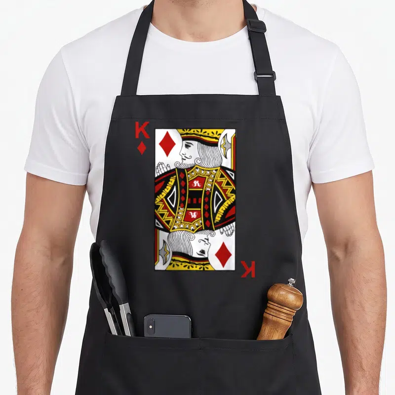 Diamond king apron - Cartoon King of Diamonds playing card with red K and diamond symbols — shown on a black cotton canvas apron against a white background