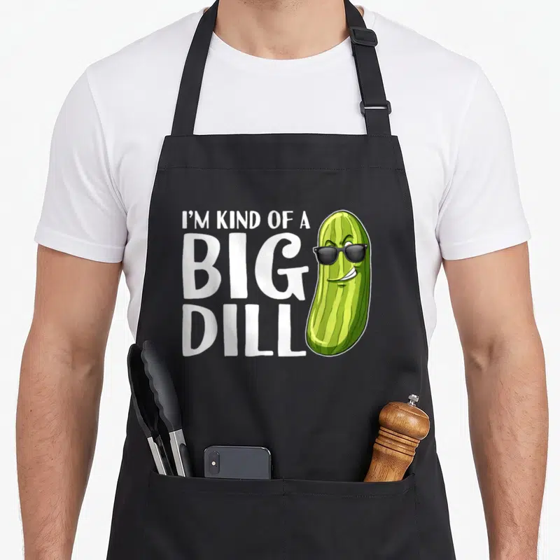 Kind pickle apron - Cartoon pickle with sunglasses next to stacked white text reading I'M KIND OF A, BIG, DILL — shown on a black cotton canvas apron against a white background