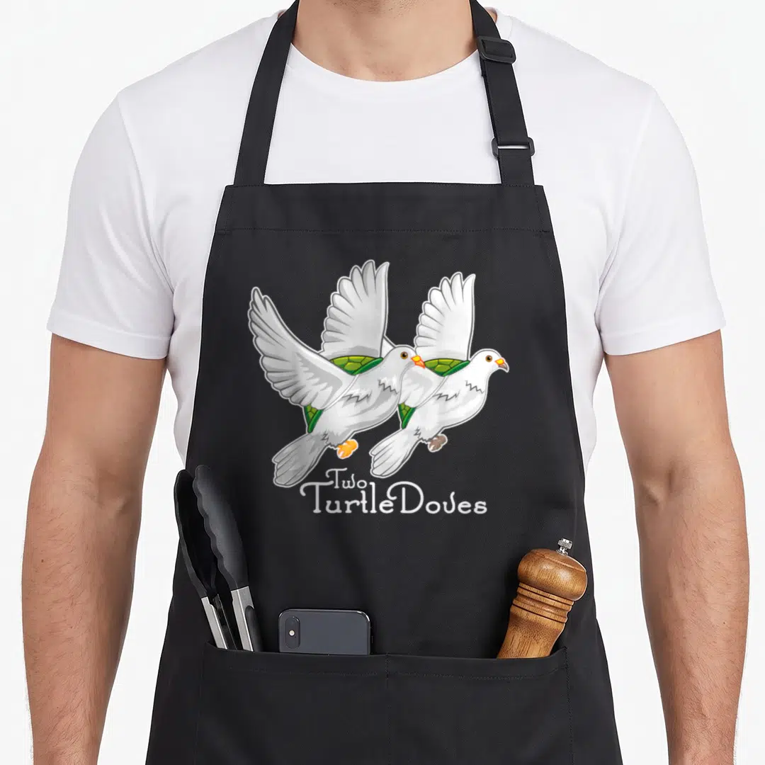 Turtle christmas apron - Two white cartoon doves flying with green turtle shells on backs and text Two Turtle Doves — shown on a black cotton canvas apron against a white background