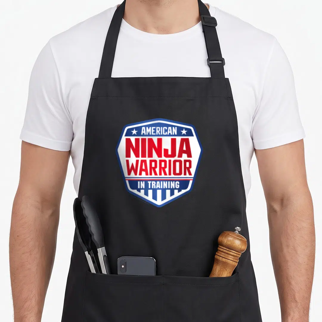 Ninja training apron - Shield logo with AMERICAN NINJA WARRIOR IN TRAINING text and stars — shown on a black cotton canvas apron against a white background