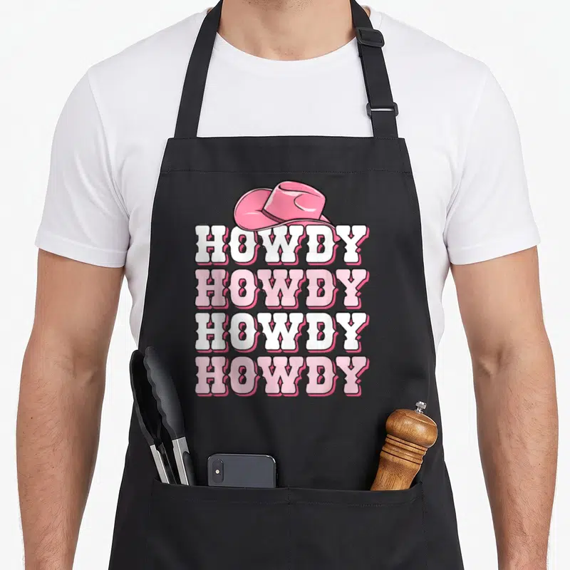 Howdy western aprons - Pink cowboy hat above four stacked HOWDY words in white and pink cartoon font — shown on a black cotton canvas apron against a white background