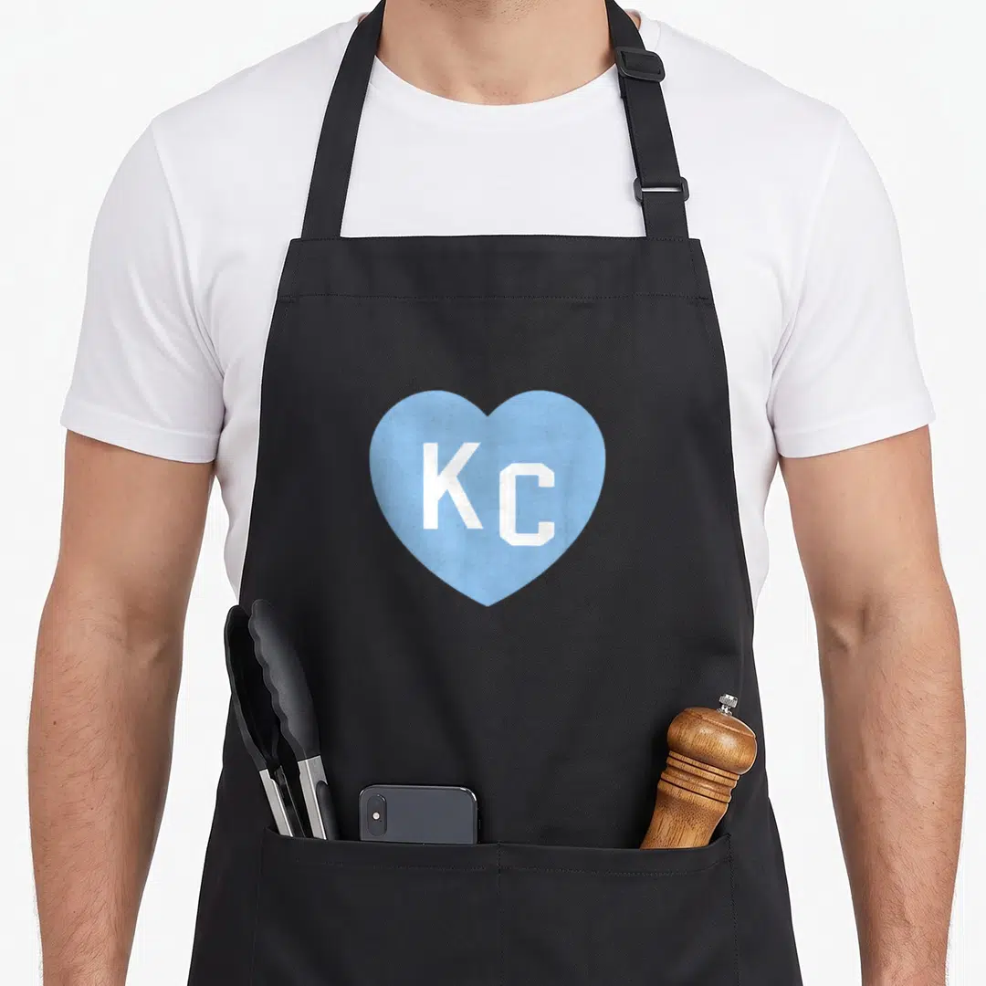 Letter kansas apron - Light blue heart containing white KC letters centered on gray background — shown on a black cotton canvas apron against a white background