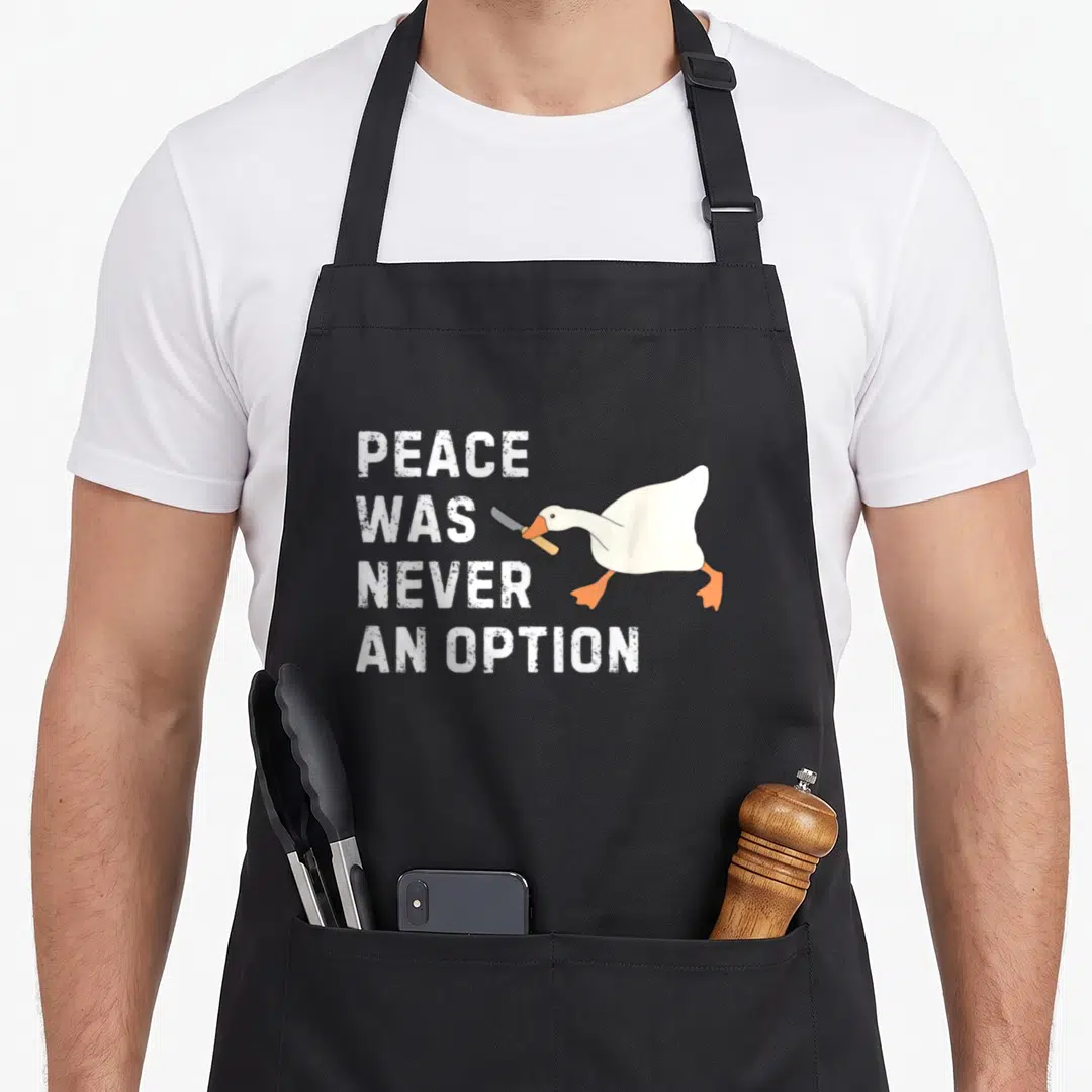 Peace option goose apron - White cartoon goose holding knife next to stacked white text on grey background — shown on a black cotton canvas apron against a white background