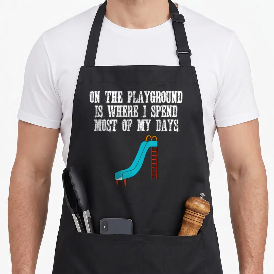 Vintage spend playground apron - White stacked text above blue slide with red ladder and yellow rails — shown on a black cotton canvas apron against a white background