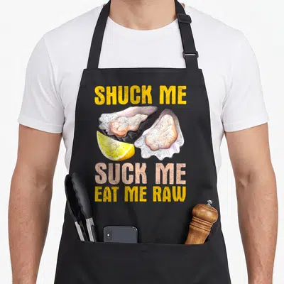 Suck oyster apron - Two open oysters and lemon slice surrounded by SHUCK ME SUCK ME EAT ME RAW text — shown on a black cotton canvas apron against a white background