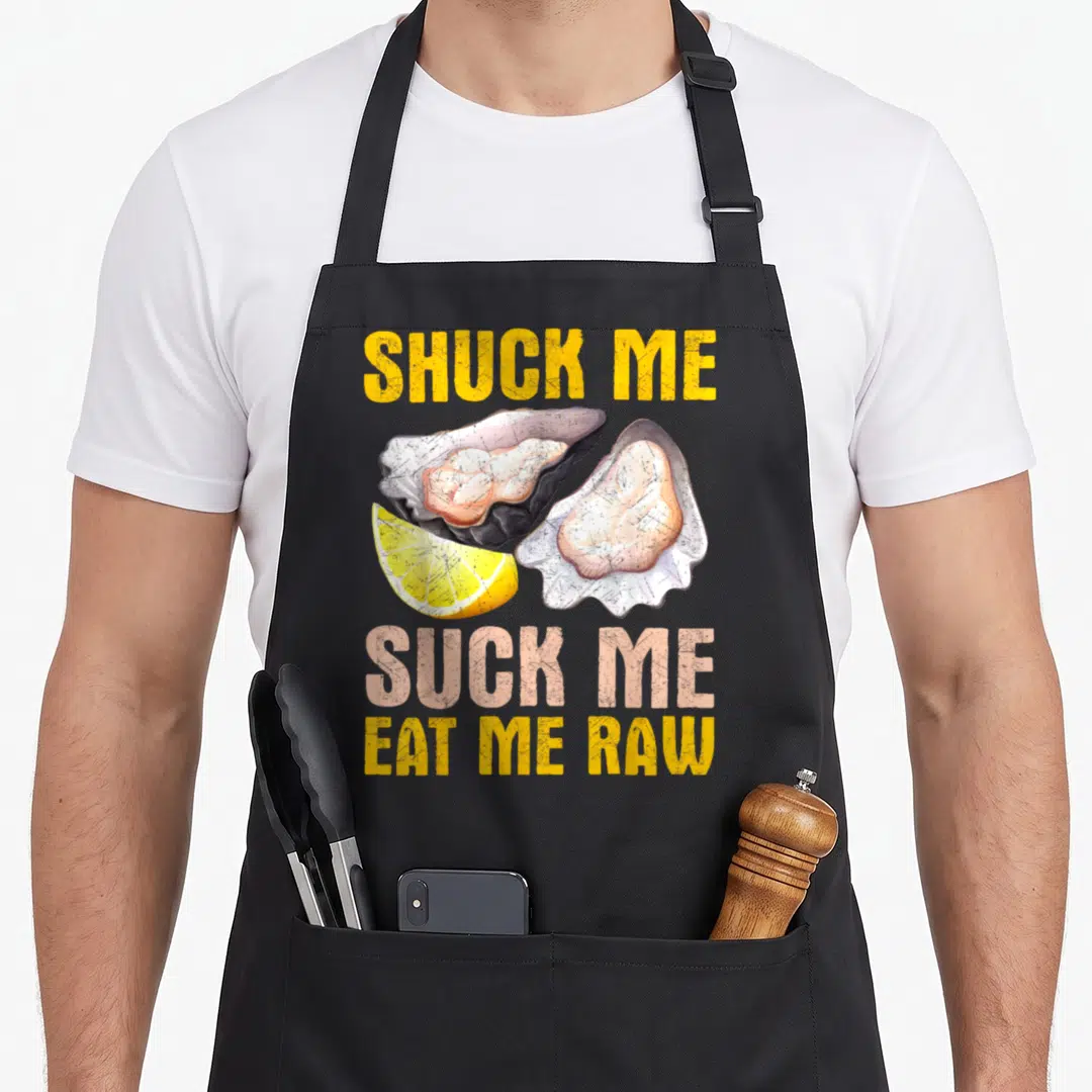 Suck oyster apron - Two open oysters and lemon slice surrounded by SHUCK ME SUCK ME EAT ME RAW text — shown on a black cotton canvas apron against a white background