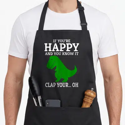 Funny clap t-rex apron - Green T-Rex silhouette with white text IF YOU'RE HAPPY AND YOU KNOW IT CLAP YOUR OH — shown on a black cotton canvas apron against a white background