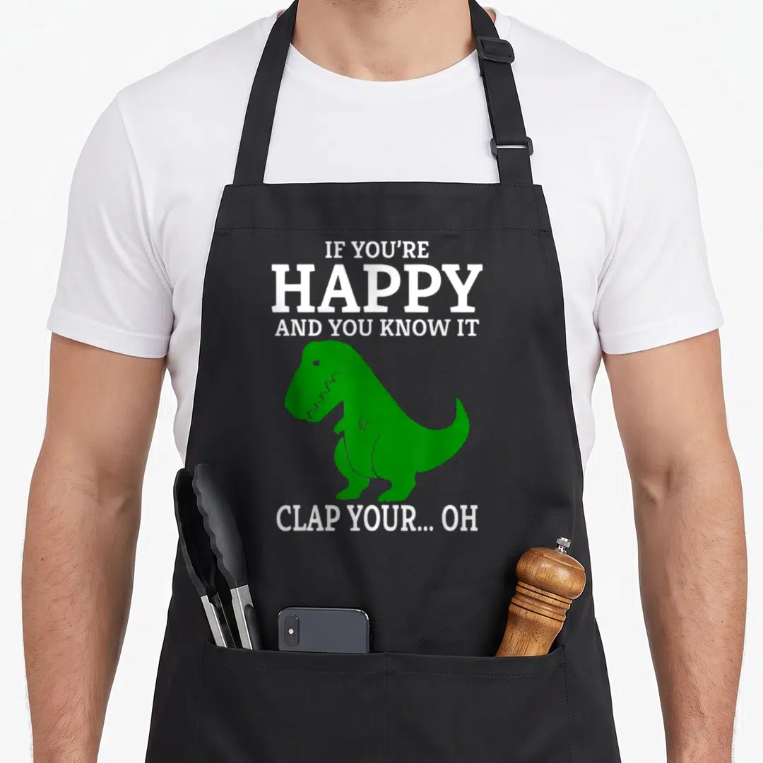 Funny clap t-rex apron - Green T-Rex silhouette with white text IF YOU'RE HAPPY AND YOU KNOW IT CLAP YOUR OH — shown on a black cotton canvas apron against a white background