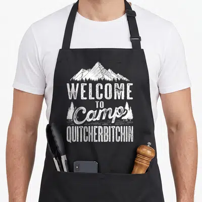Quitcherbitchin camping apron - White distressed mountain graphic and sarcastic camp text on black bib — shown on a black cotton canvas apron against a white background