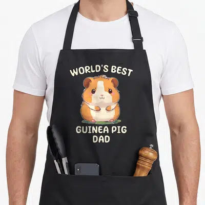 Guinea papa apron - Cartoon orange and white guinea pig sitting on green leaves under arched text reading World's Best — shown on a black cotton canvas apron against a white background