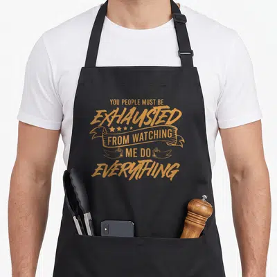 Vintage must exhausted apron - Orange stacked text and ribbon banners reading You People Must Be Exhausted — shown on a black cotton canvas apron against a white background