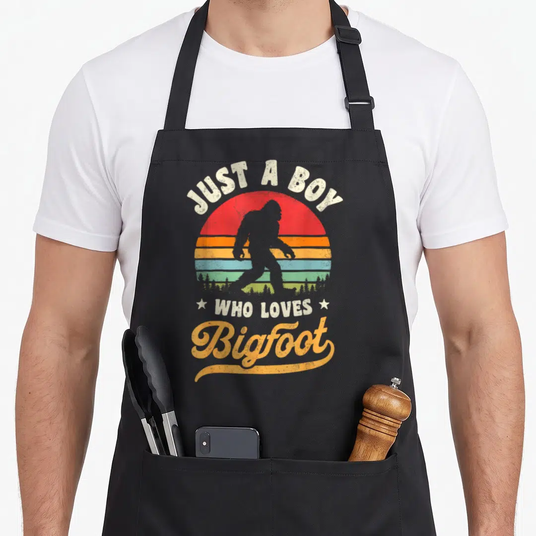 Cute sasquatch bigfoot apron - Retro striped sun with Bigfoot silhouette and Just A Boy Who Loves Bigfoot text — shown on a black cotton canvas apron against a white background