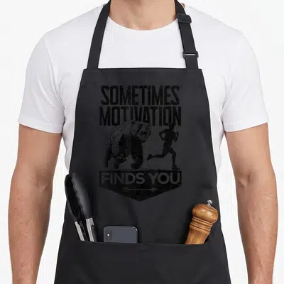 Sometime find aprons - Distressed black bear chasing runner with text Sometimes Motivation Finds You — shown on a black cotton canvas apron against a white background