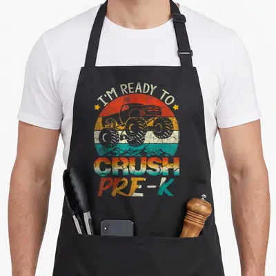 Crush pre truck apron - Retro monster truck in red orange yellow teal sun with text I'M READY TO CRUSH PRE-K — shown on a black cotton canvas apron against a white background