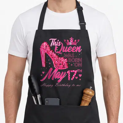 17th heel birthday apron - Pink high heel shoe with crown and May 17th birthday text — shown on a black cotton canvas apron against a white background