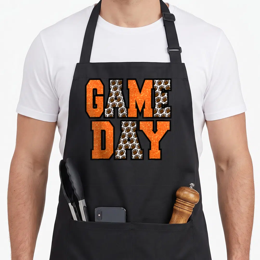 Cute school football apron - GAME DAY text in orange glitter with brown football pattern filling the letter — shown on a black cotton canvas apron against a white background