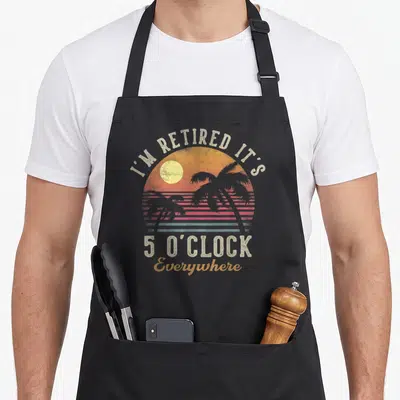 Oclock retirement apron - Retro sunset circle with palm trees and text I'M RETIRED IT'S 5 O'CLOCK Everywhere in beige — shown on a black cotton canvas apron against a white background