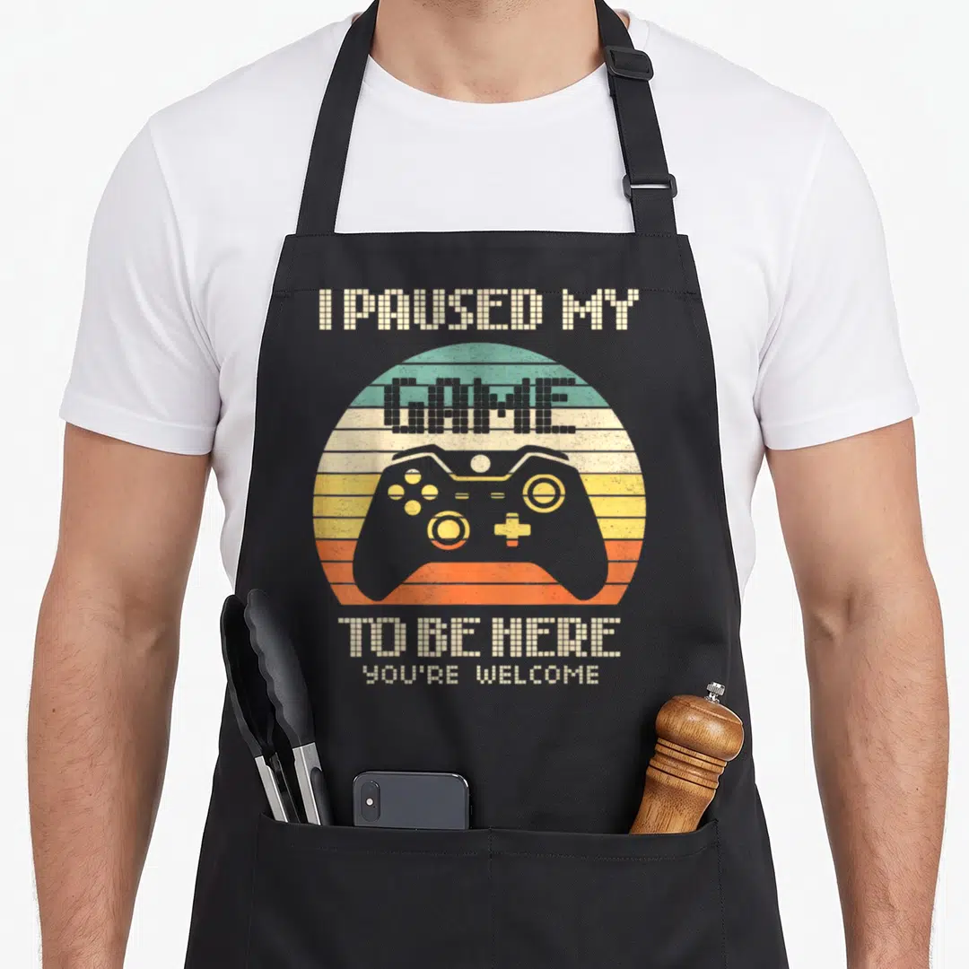 Paused game aprons - Grey controller on teal orange and yellow stripes with pixelated text — shown on a black cotton canvas apron against a white background