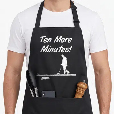Ten metal detecting apron - White Ten More Minutes text above curler silhouette with broom on ice strip — shown on a black cotton canvas apron against a white background