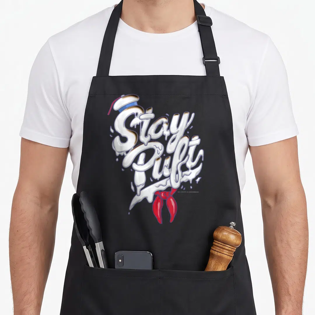 Font stay puft apron - Stay Puft text styled as melting marshmallows with sailor hat and red tie in white blue and red — shown on a black cotton canvas apron against a white background