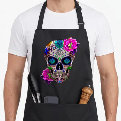 Sugar day of the dead apron - Cartoon sugar skull with pink roses, purple leaves, and blue eye details on black fabric — shown on a black cotton canvas apron against a white background