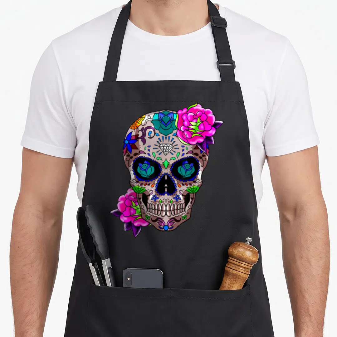 Sugar day of the dead apron - Cartoon sugar skull with pink roses, purple leaves, and blue eye details on black fabric — shown on a black cotton canvas apron against a white background