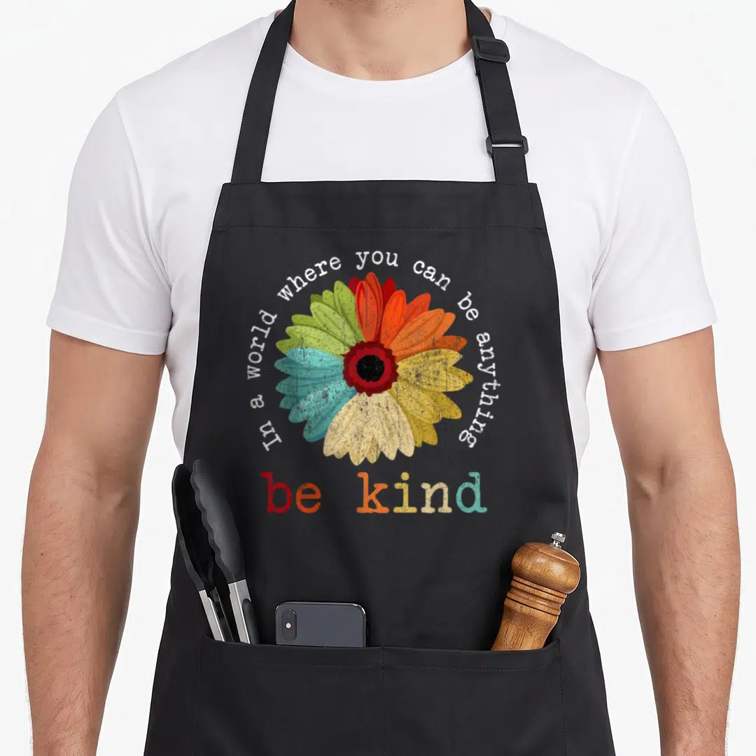 Kindness novelty apron - Multicolored cartoon flower with arched text reading in a world where you can be anything be — shown on a black cotton canvas apron against a white background