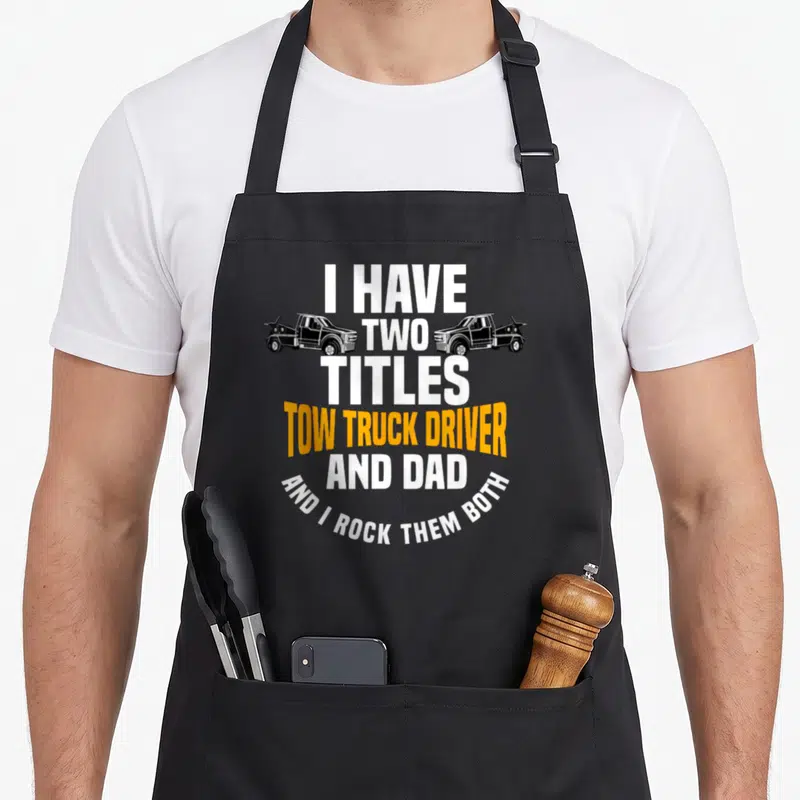 Vehicle father day apron - Stacked text I have two titles tow truck driver and dad with two black tow trucks facing — shown on a black cotton canvas apron against a white background