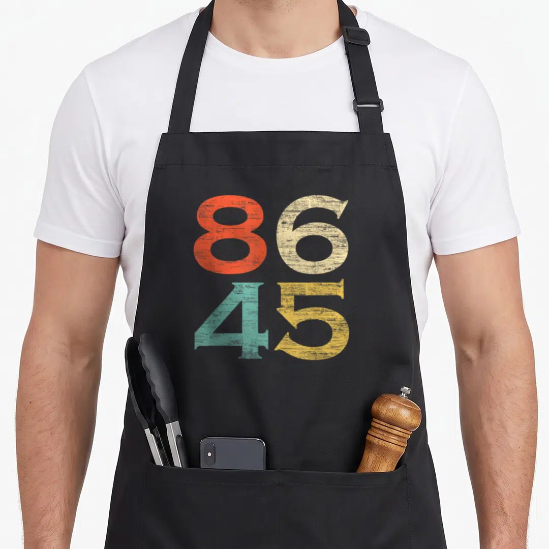 Anti birthday mode apron - Stacked 86 and 45 numbers in red beige teal and yellow distressed font — shown on a black cotton canvas apron against a white background