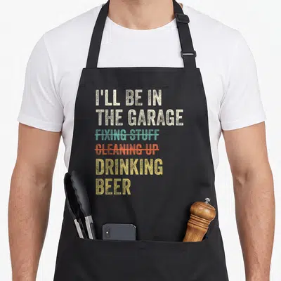 Drinking garage apron - Stacked beige teal orange yellow distressed text with strike-through lines reading I'll be — shown on a black cotton canvas apron against a white background