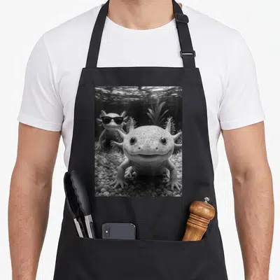 Axolotl selfie apron - Two axolotls on pebbles, one wearing sunglasses in grayscale photo style — shown on a black cotton canvas apron against a white background