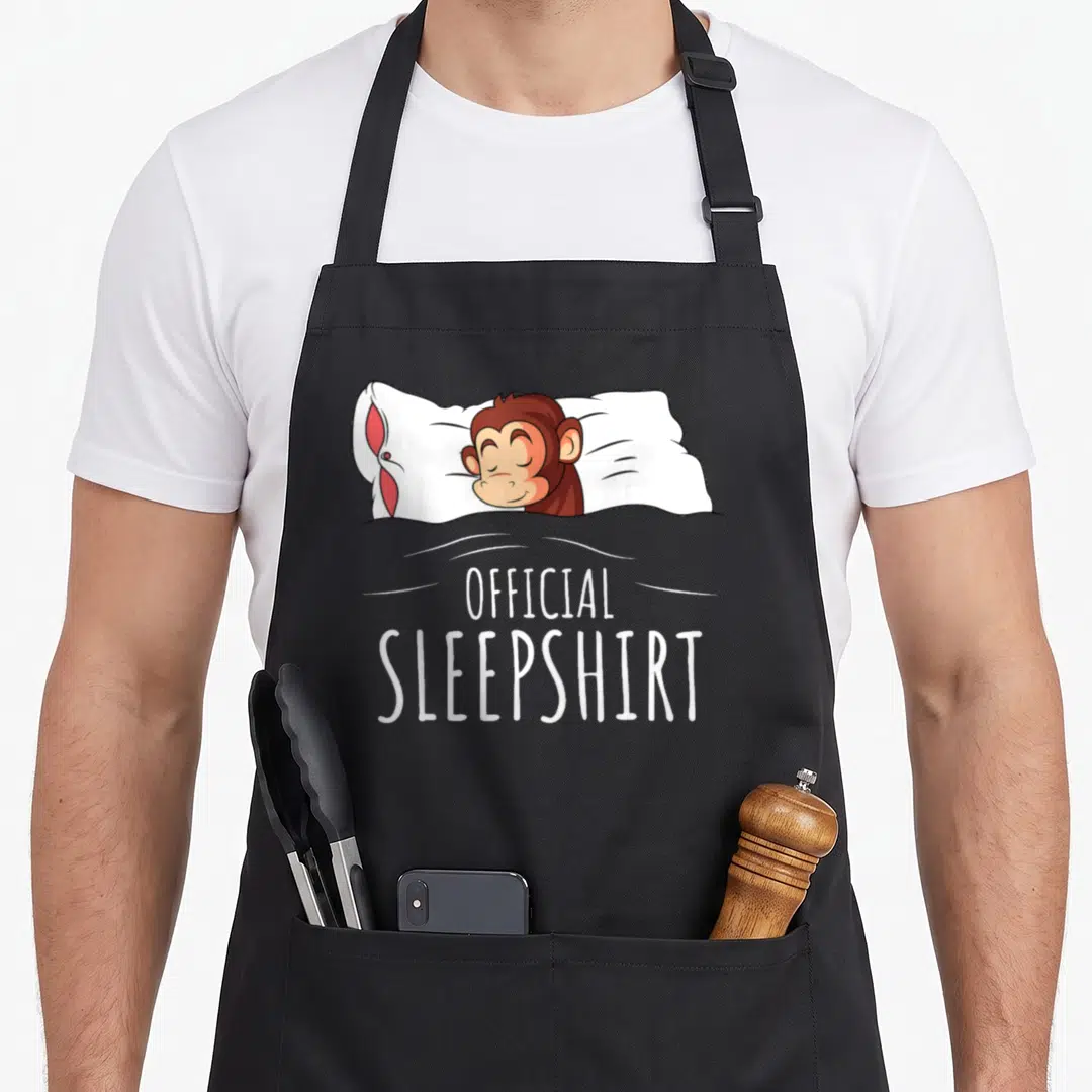 Monkey novelty apron - Cartoon brown monkey sleeping on white pillow with red details under grey blanket above white — shown on a black cotton canvas apron against a white background