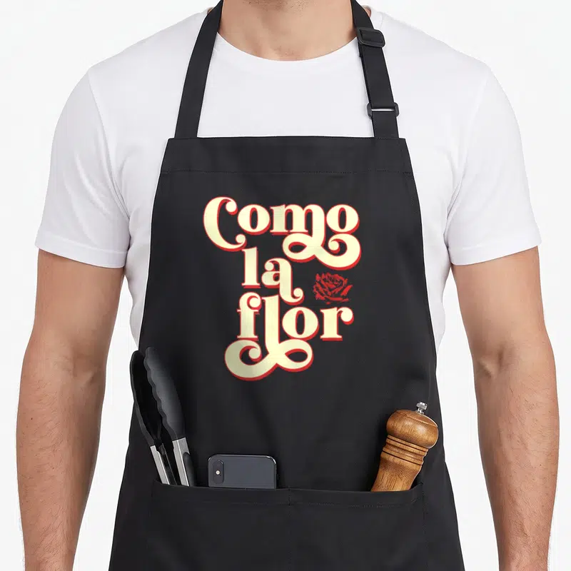 Cinco modern apron - Stacked Como la flor text in cream and red with a small rose icon — shown on a black cotton canvas apron against a white background