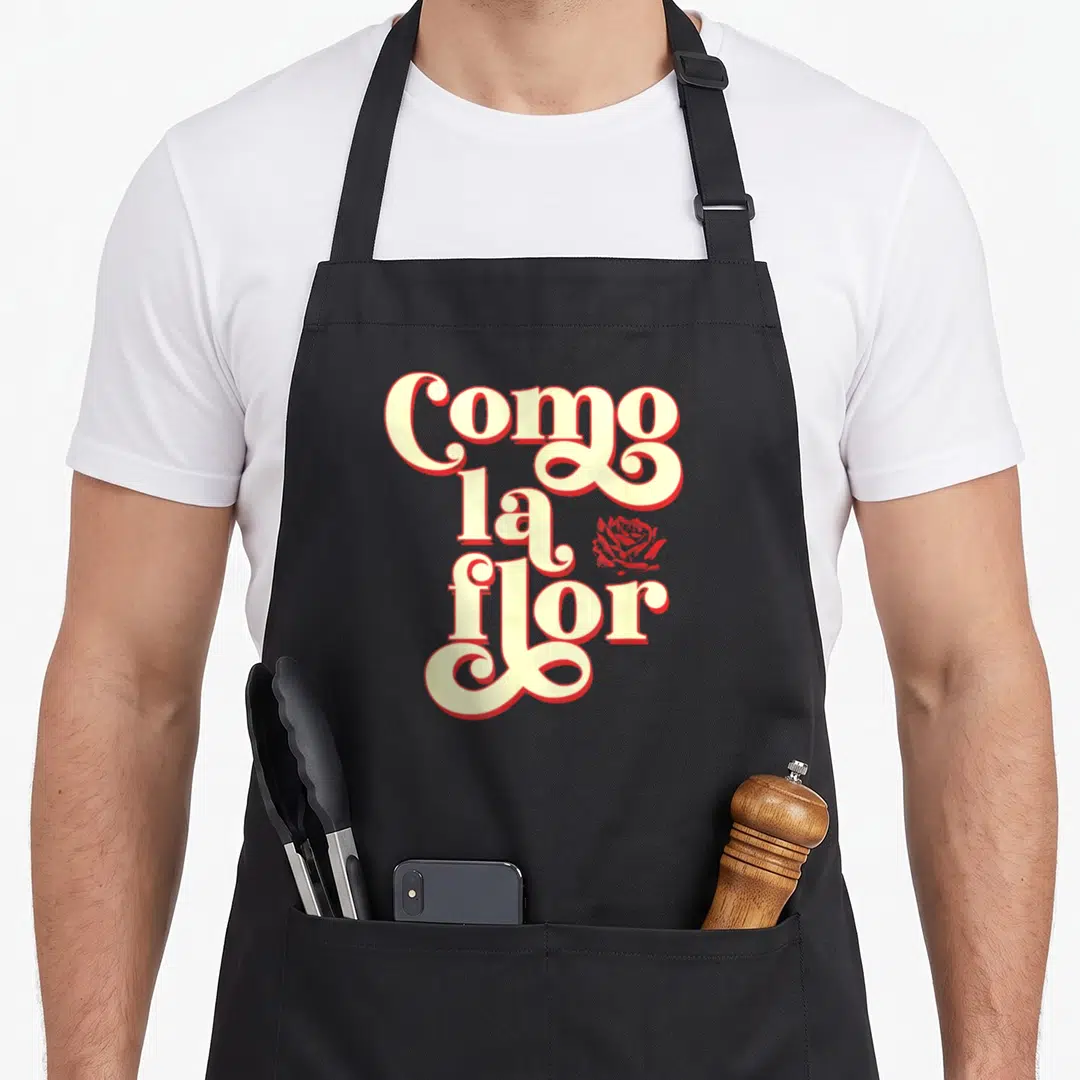 Cinco modern apron - Stacked Como la flor text in cream and red with a small rose icon — shown on a black cotton canvas apron against a white background