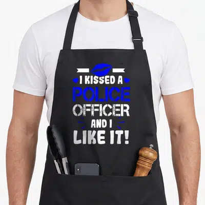 Kissed police wife apron - Blue lips on white ribbon above stacked text reading I kissed a police officer and I like it — shown on a black cotton canvas apron against a white background