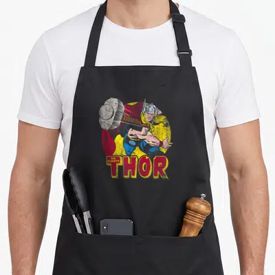 Comic hammer apron - Thor holding grey hammer in red blue yellow retro comic with stacked THE MIGHTY THOR text below — shown on a black cotton canvas apron against a white background