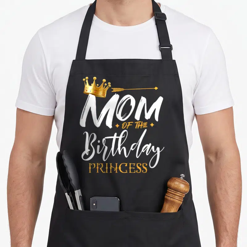 Birthday grandma apron - Gold crown above white MOM OF THE Birthday PRINCESS text with orange arrow — shown on a black cotton canvas apron against a white background