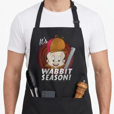 Elmer looney tune apron - Elmer Fudd head over red target rings with white It's WABBIT SEASON text — shown on a black cotton canvas apron against a white background