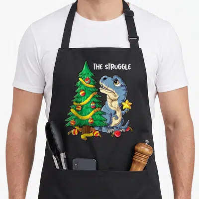 Rex christmas tree apron - Blue dinosaur holding star next to green tree with red ornaments and THE STRUGGLE text — shown on a black cotton canvas apron against a white background