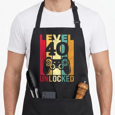 Level unlocked 40th apron - Grey game controller over five vertical stripes in red, yellow, teal, orange, and grey — shown on a black cotton canvas apron against a white background