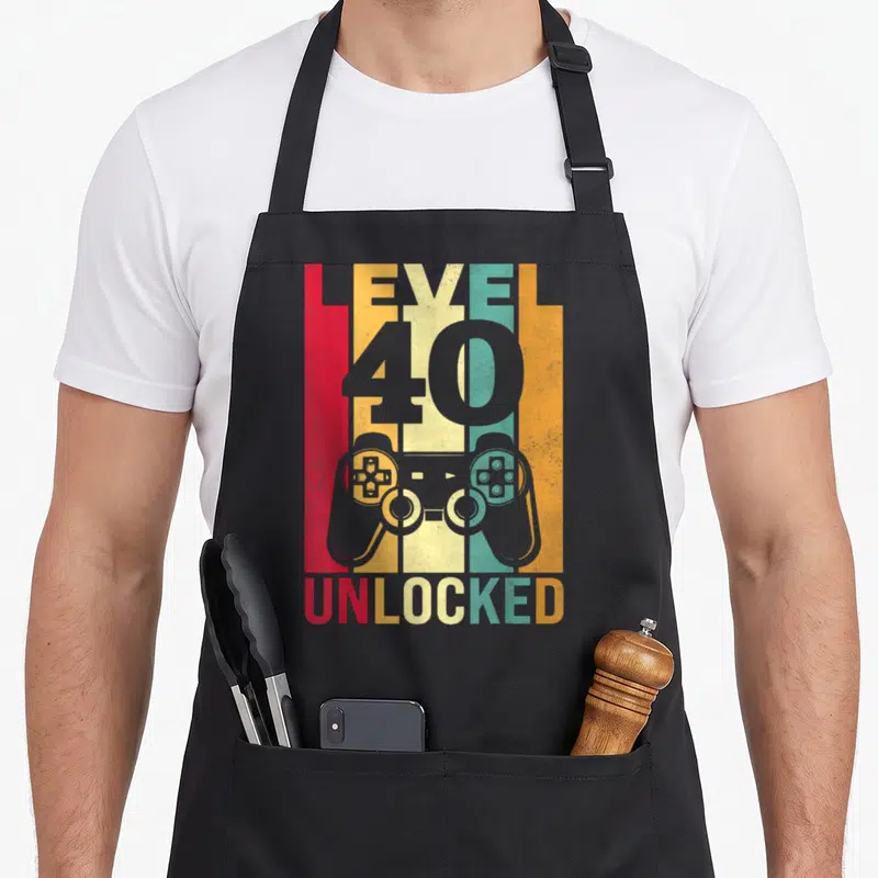 Level unlocked 40th apron - Grey game controller over five vertical stripes in red, yellow, teal, orange, and grey — shown on a black cotton canvas apron against a white background