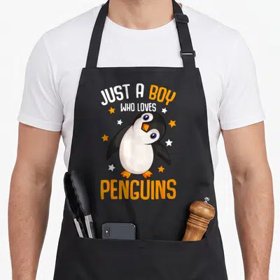 Penguin aprons gift - Cartoon penguin with orange beak and stars between text Just a Boy Who Loves Penguins — shown on a black cotton canvas apron against a white background
