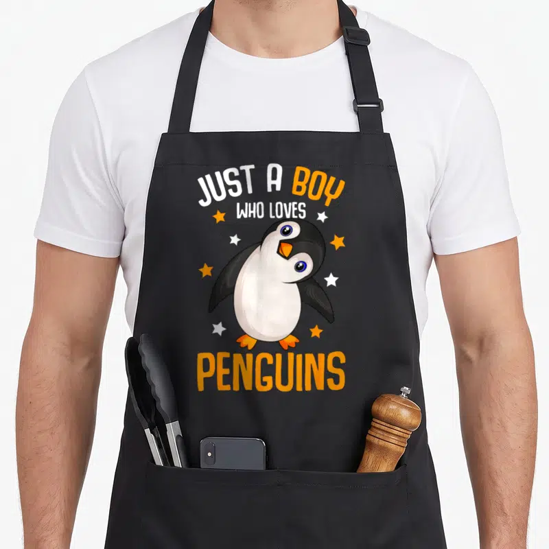Penguin aprons gift - Cartoon penguin with orange beak and stars between text Just a Boy Who Loves Penguins — shown on a black cotton canvas apron against a white background