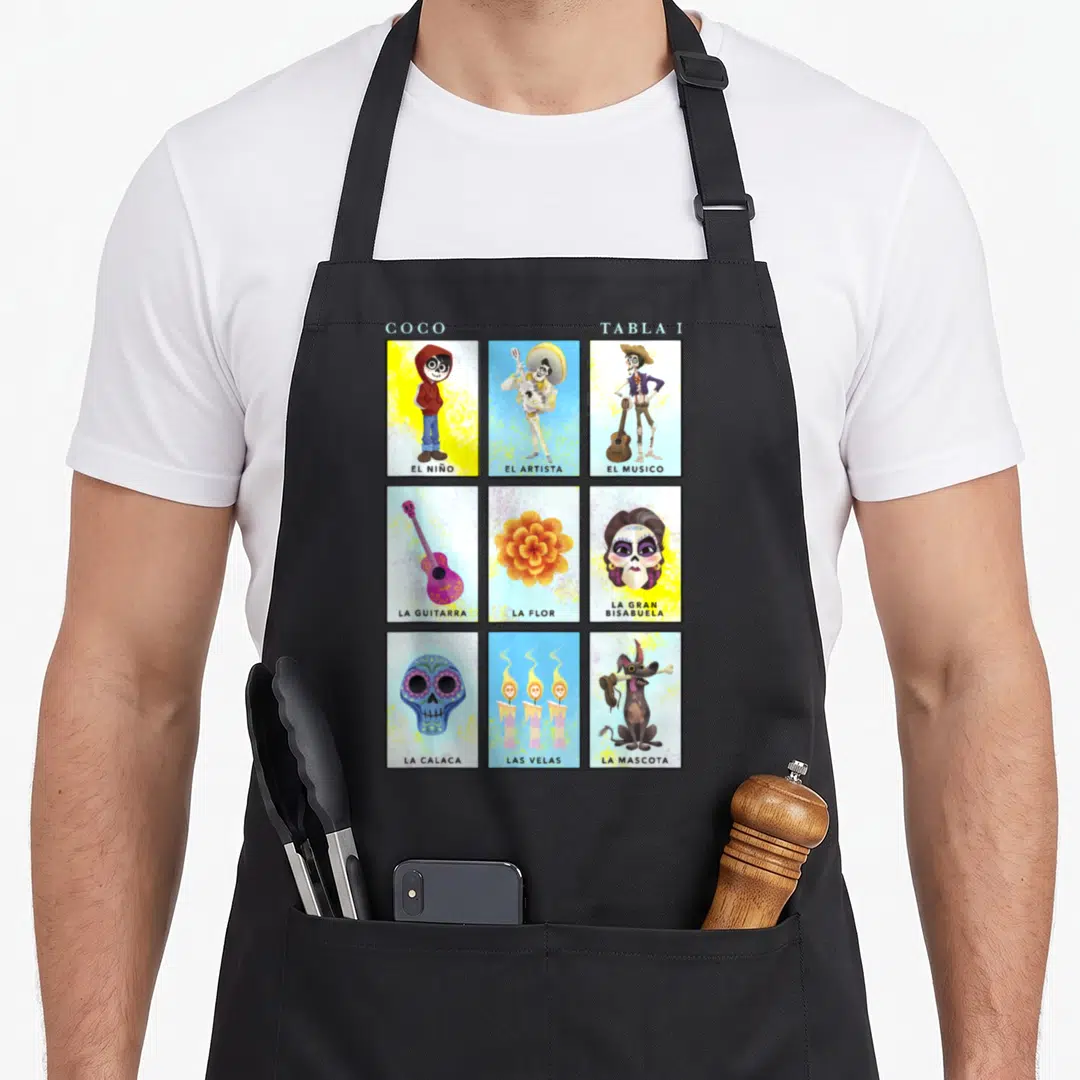 Coco table apron - Nine cartoon panels showing El Niño, La Flor, and La Calaca with Spanish labels — shown on a black cotton canvas apron against a white background