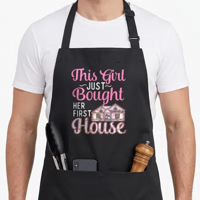 Bought homeowner housewarming apron - Pink and white text reading This Girl Just Bought Her First House with floral — shown on a black cotton canvas apron against a white background