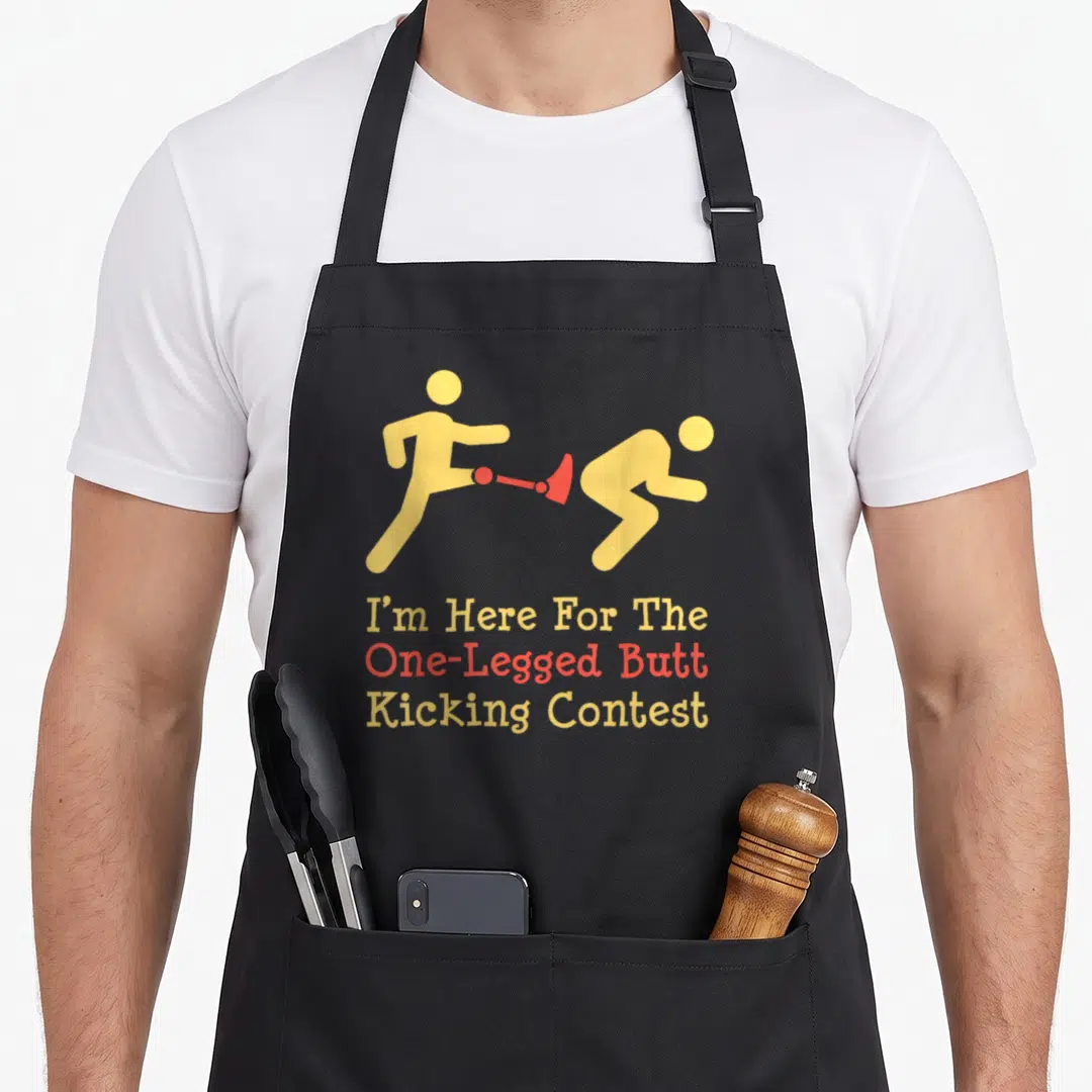 Legged leg apron - Two yellow figures kicking with red prosthetic leg above stacked contest text — shown on a black cotton canvas apron against a white background