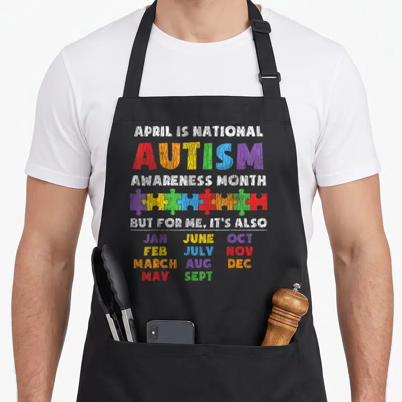 National autism awareness apron - Multi-colored distressed text listing all months with a row of interlocking puzzle — shown on a black cotton canvas apron against a white background