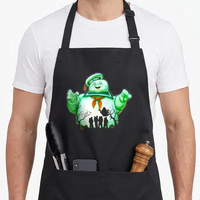 Shot marshmallow apron - Green Stay Puft Marshmallow Man with internal silhouette of four people, trees, pumpkins — shown on a black cotton canvas apron against a white background