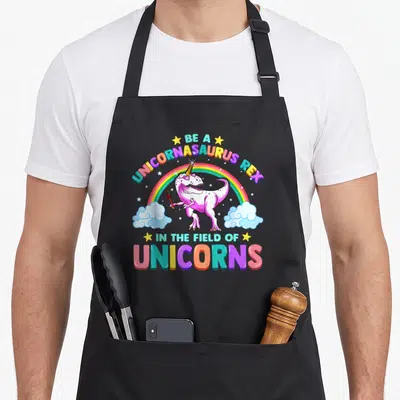 Unicornasaurus unicorn apron - Pink dinosaur holding wand under rainbow with stars and arched text — shown on a black cotton canvas apron against a white background